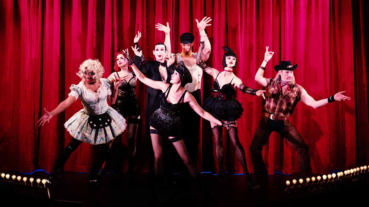 Tim Fischer & Ensemble in schwarzen und weißen Burlesque-Kostümen vor rotem Bühnenvorhang, posierend mit ausgestreckten Armen.