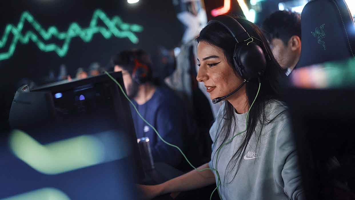 Drei Personen sitzen an Computern mit Gaming-Stühlen, eine trägt ein graues Nike-Sweatshirt und ein Headset, im Hintergrund grüne Linien auf dunklem Bildschirm.