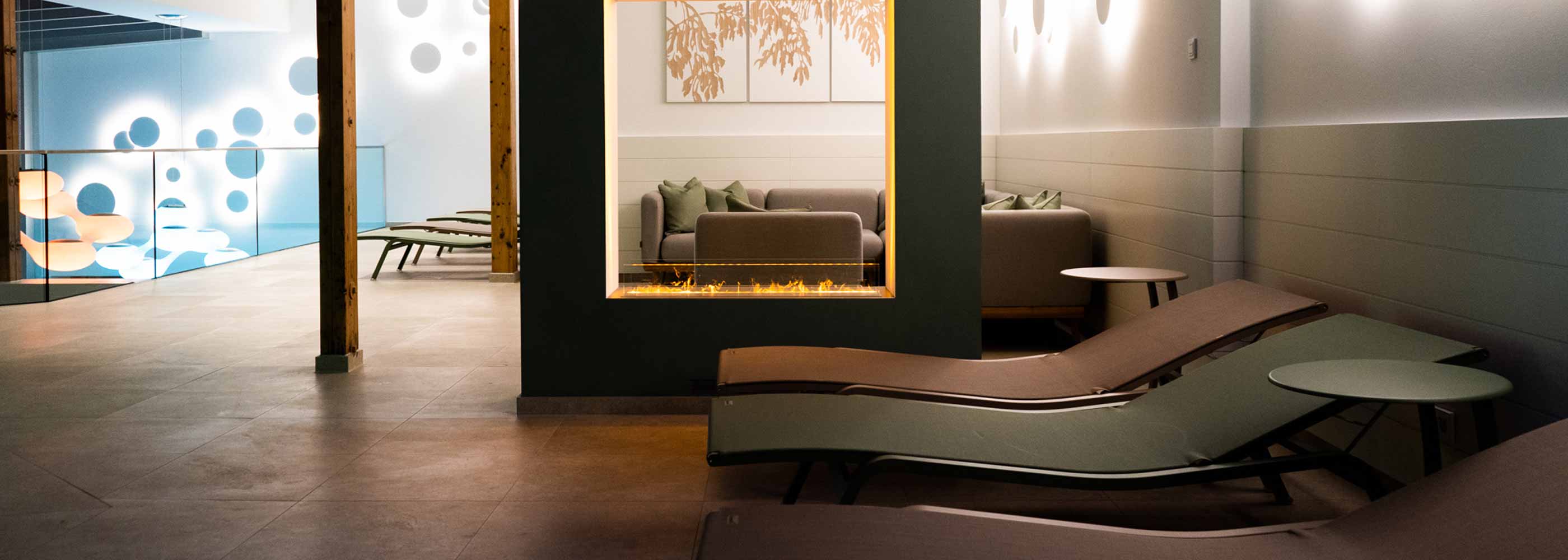 Moderner Ruheraum mit zwei braunen Liegen, einem eingebauten Kamin und einem Sitzbereich mit grauem Sofa im Hintergrund.