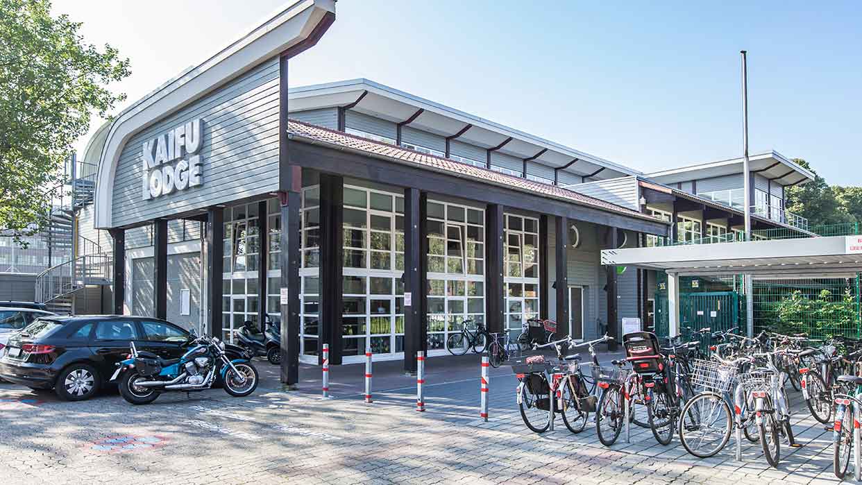 Ansicht der KAIFU LODGE, ein modernes Gebäude mit großen Fenstern und einem Vordach. Vor dem Gebäude stehen mehrere Fahrräder und Autos.