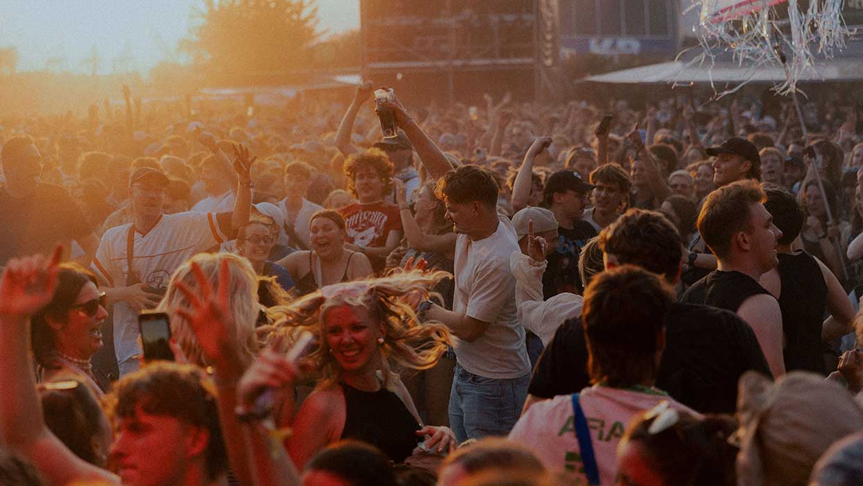 Menschenmenge bei Sonnenuntergang auf einem Festival, viele mit erhobenen Armen und lässiger Kleidung.