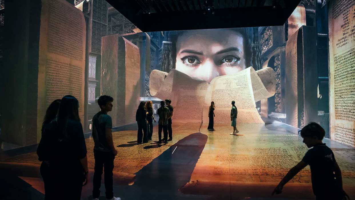 Menschen stehen in einem großen, von warmem Licht durchfluteten Raum mit hohen, steinernen Säulen und Wänden, die mit Texten bedeckt sind.