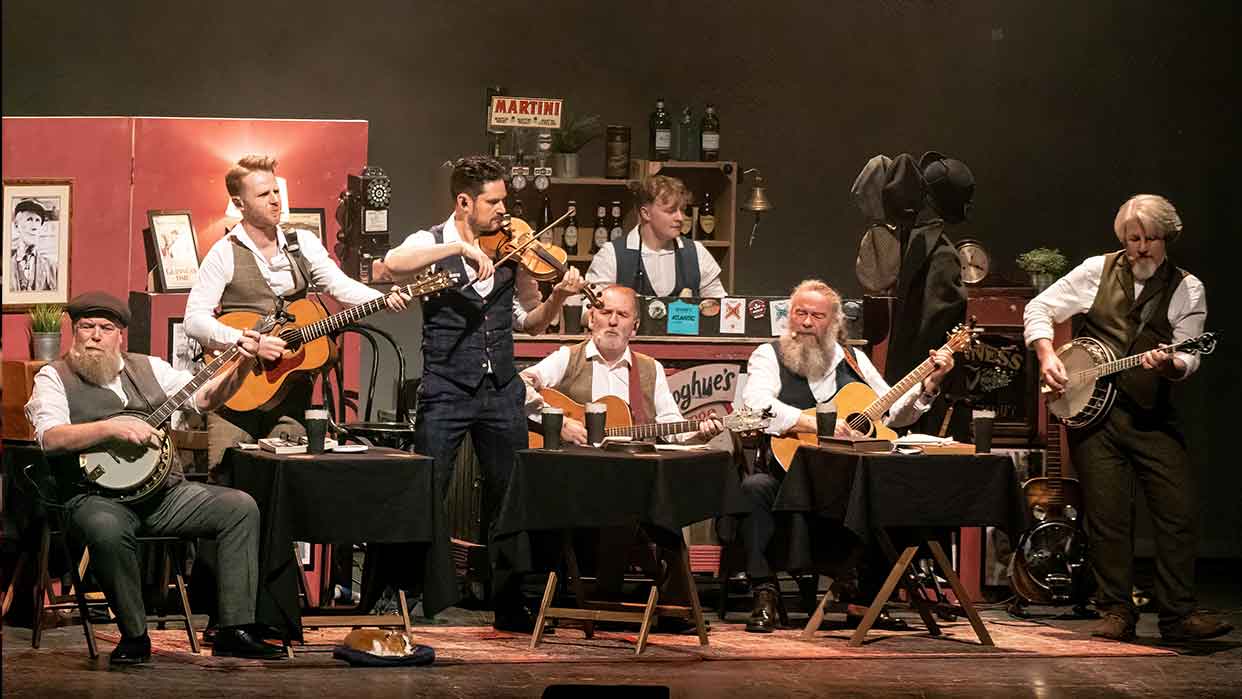 Musikgruppe mit sieben Mitgliedern spielt auf einer Bühne. Sie halten verschiedene Instrumente wie Gitarre, Banjo und Violine. 