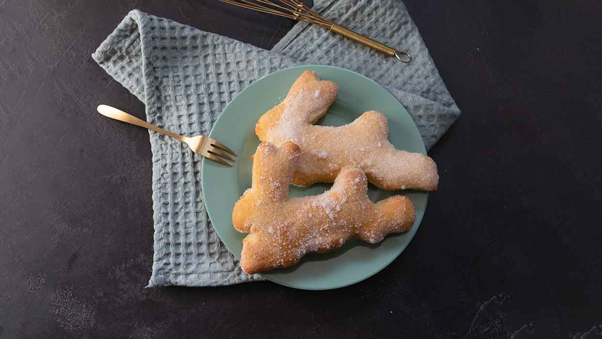 Zwei zu Hasen geformte Quarkteiggebäcke auf einem grünen Teller mit goldfarbenem Besteck auf grauem Stoff.