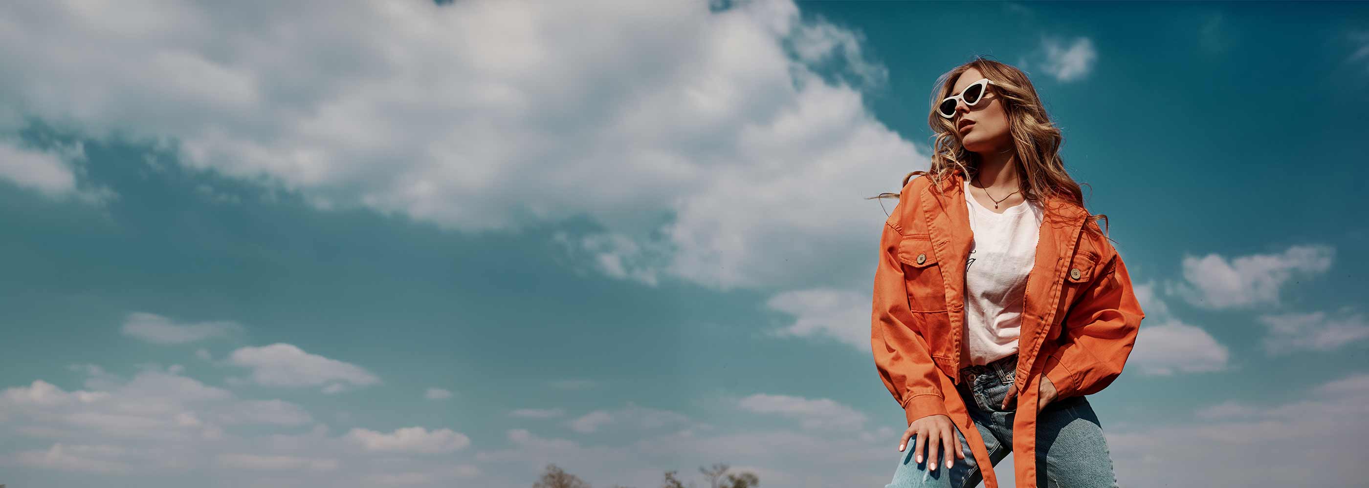 Person mit orangefarbener Jacke und weißem T-Shirt steht vor blauem Himmel mit Wolken.