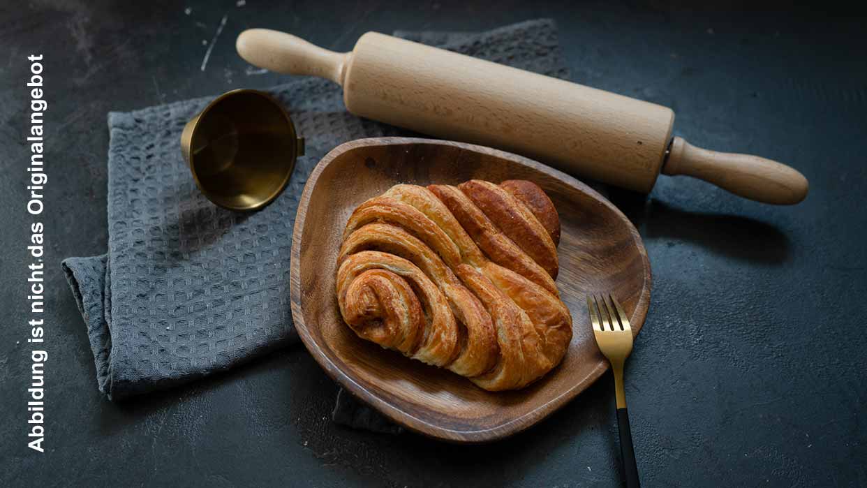 Ein frisch gebackenes Franzbrötchen liegt auf einem Holzteller, der auf einem Tisch platziert ist. Der Tisch ist mit einer Nudelrolle und einem Küchentuch dekoriert.