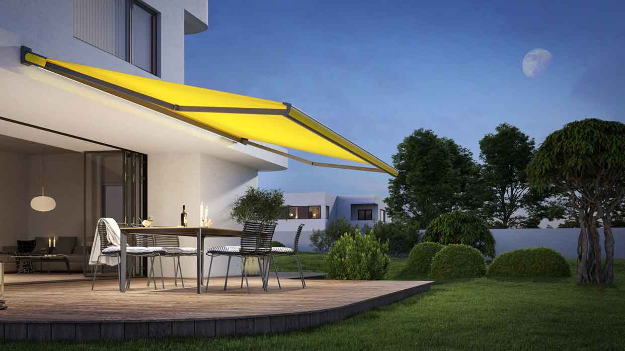 Terrasse mit gelber Markise, Tisch mit Stühlen und Weinflasche vor modernem Haus im Garten bei Abenddämmerung.