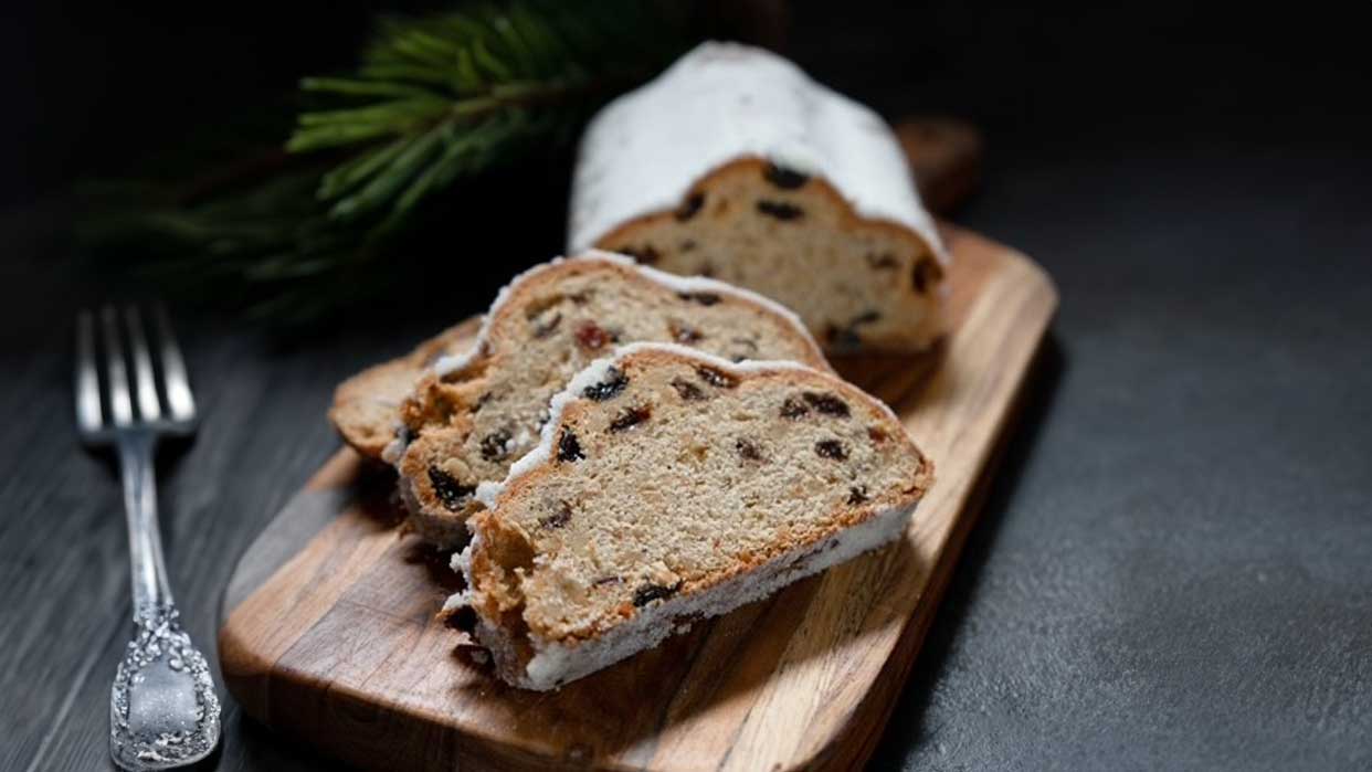 Aufgeschnittener Christstollen mit Puderzucker und Rosinen auf Holzbrett, daneben eine Gabel und Tannenzweige.