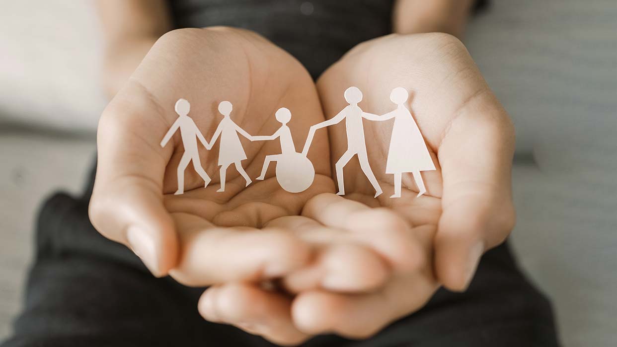 Zwei Hände halten ausgeschnittene Papierfiguren, die eine vielfältige Familie darstellen – darunter Erwachsene, Kinder und eine Figur in einem Rollstuhl in der Mitte. Das Bild symbolisiert Inklusion, Zusammenhalt und Vielfalt.