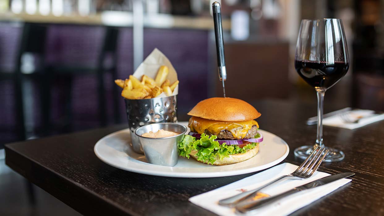 Burger mit Salat, Zwiebeln und Käse auf einem weißen Teller, daneben Pommes in einem Metallbehälter und ein Glas Rotwein auf einem dunklen Tisch im Hard Rock Cafe.