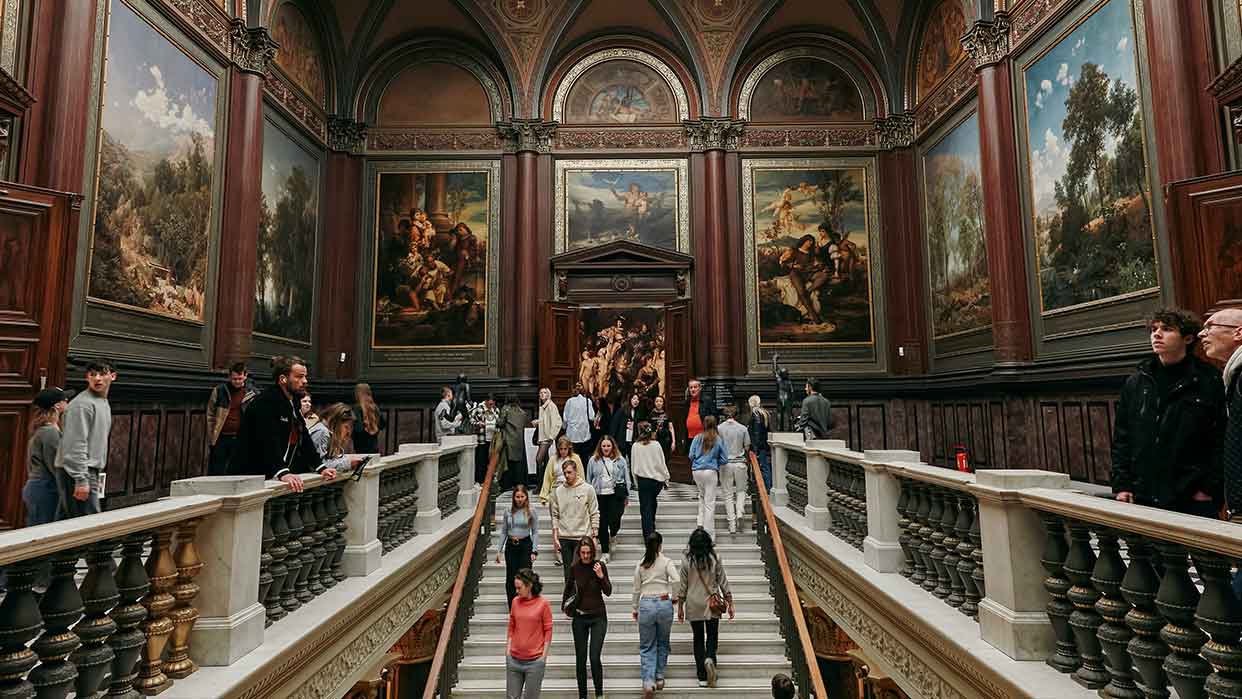Großer Museumsraum mit hohen Wänden, die mit großen Gemälden geschmückt sind, und einer breiten Treppe, auf der zahlreiche Besucher gehen und stehen.
