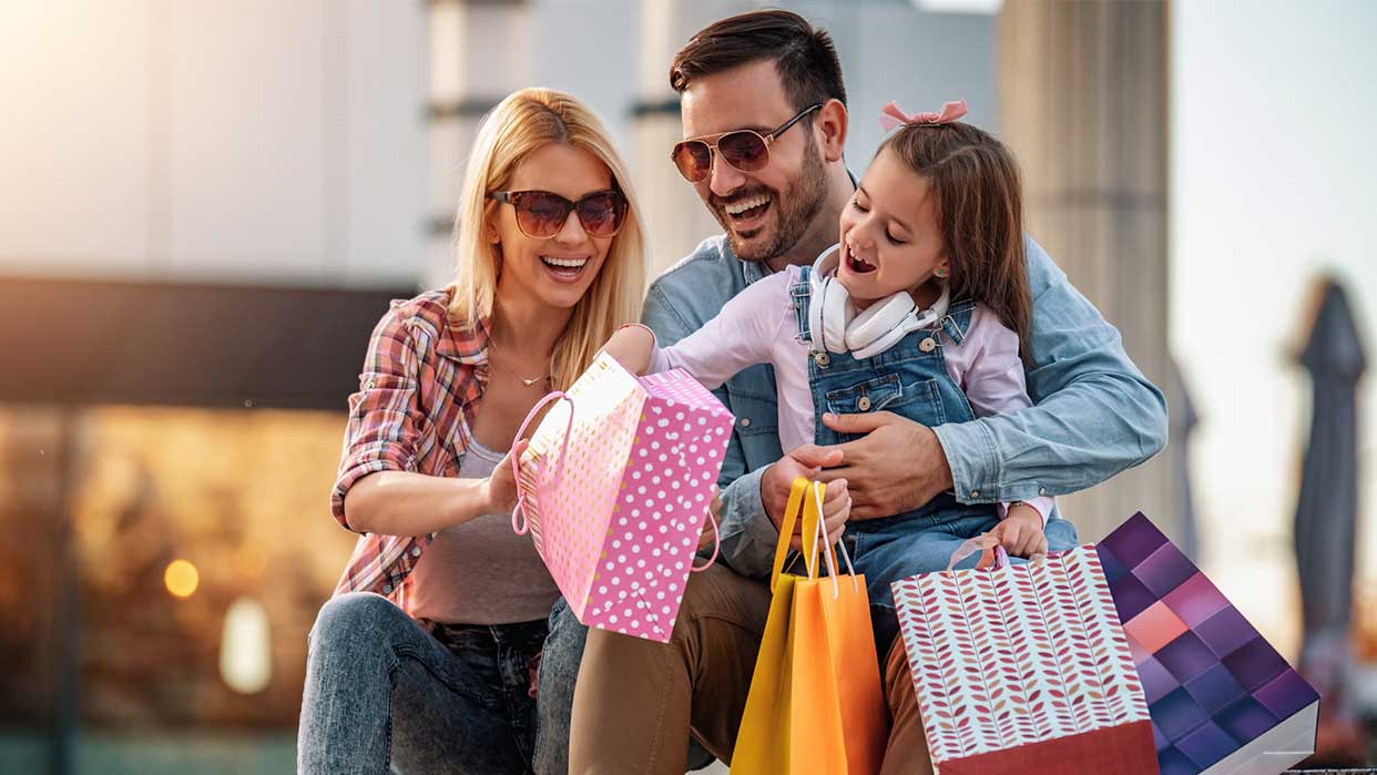 Drei Personen, eine Frau, ein Mann und ein Kind, sitzen zusammen und halten bunte Einkaufstüten mit verschiedenen Mustern.