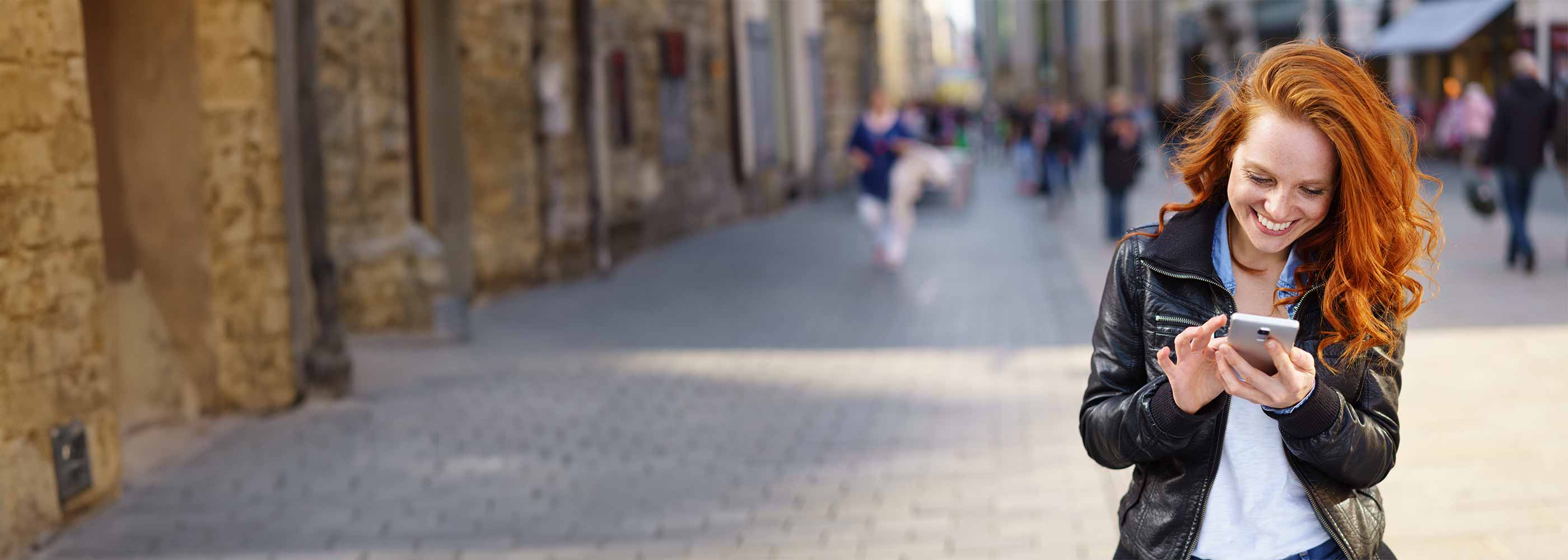 Junge Frau mit roten Haaren und Lederjacke lächelt und benutzt Smartphone auf einer belebten Straße.