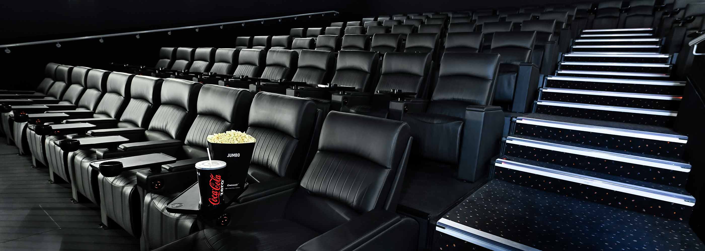 Leerer Kinosaal im CinemaxX mit schwarzen Ledersesseln und beleuchteten Treppenstufen, Popcorn und Getränk auf einem Sitz.