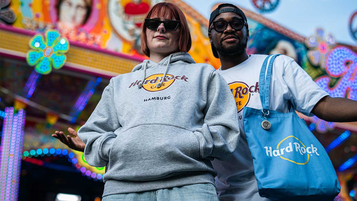 Zwei Personen vor einem bunten Hintergrund tragen Hard Rock Cafe Hamburg Kleidung: eine graue Kapuzenjacke und ein weißes T-Shirt, dazu eine blaue Hard Rock Tasche.