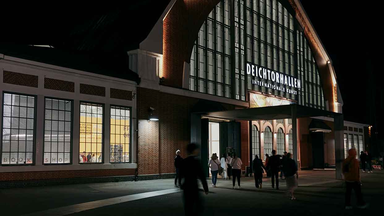 Eingang der Deichtorhallen Hamburg bei Nacht mit beleuchtetem Schriftzug und Menschen, die darauf zugehen.