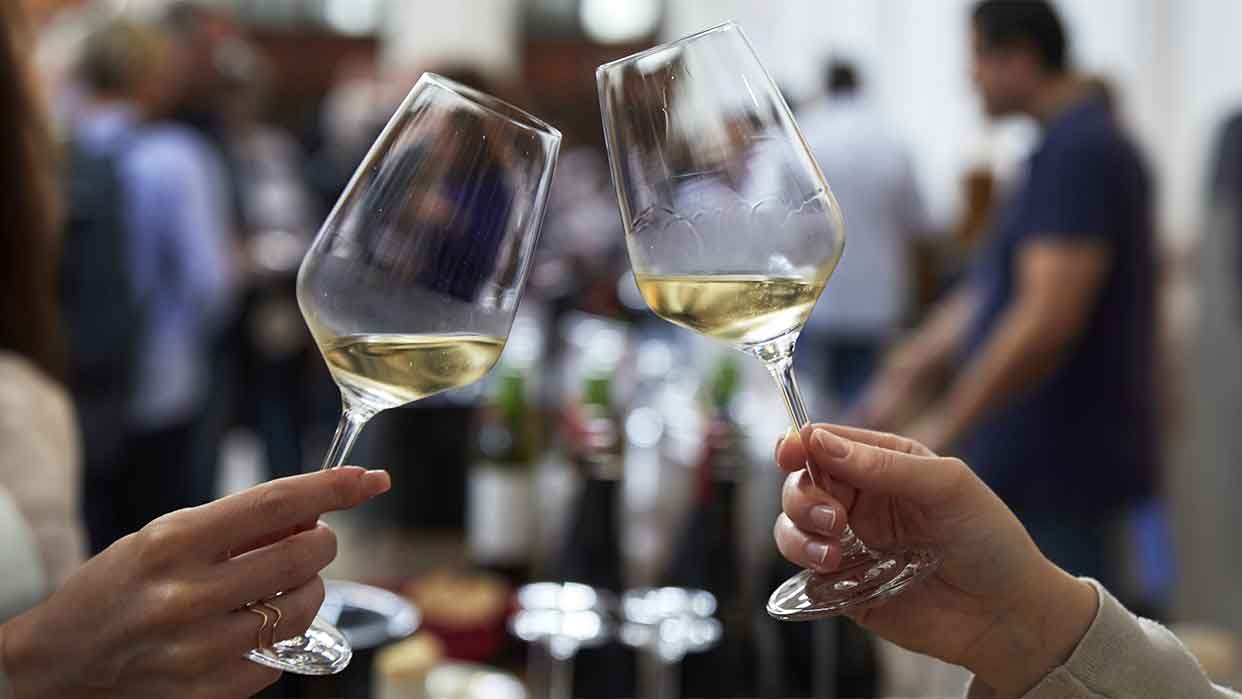 Zwei Weingläser mit Weißwein stoßen an. Im Hintergrund sind verschwommene Personen und Weinflaschen zu sehen. Event: VINORELL 2025.
