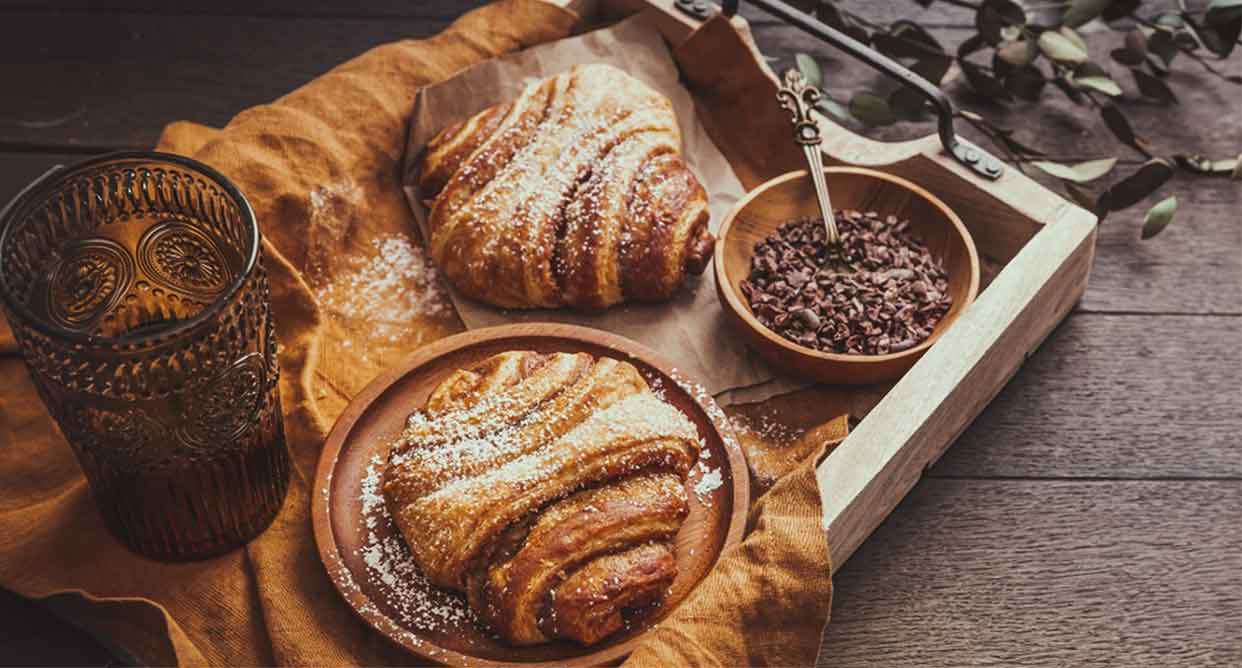 Zwei mit Puderzucker bestäubte Franzbrötchen auf braunen Tellern neben einem Glas mit Getränk und einer kleinen Schale mit Kakaonibs auf einem Holztisc