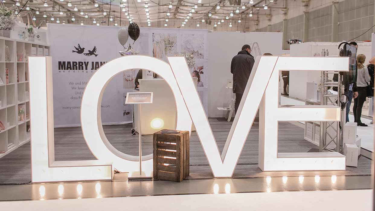 Großes beleuchtetes Schriftzug-Display mit den Buchstaben 'LOVE' auf einer Messe mit Ständen und Besuchern im Hintergrund.