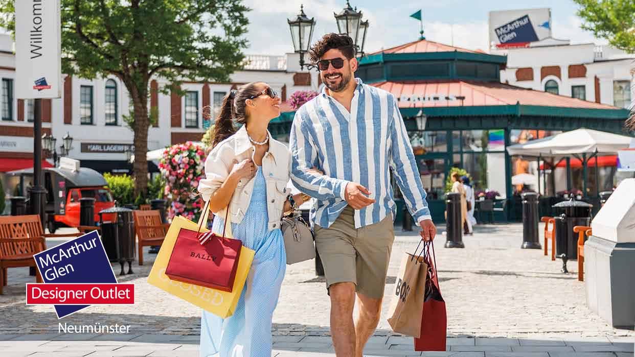 Paar geht mit Einkaufstüten durch das Designer Outlet Neumünster an einem sonnigen Tag.
