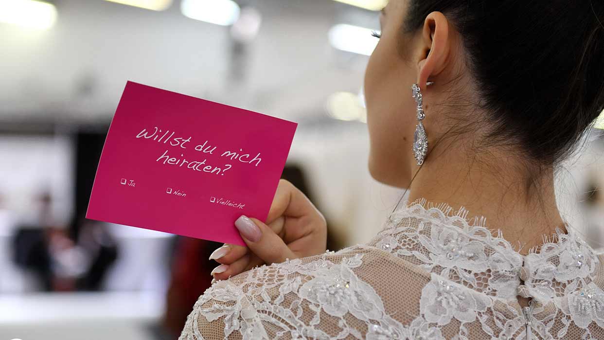 Frau in Spitzenkleid hält eine pinke Karte mit der Aufschrift 'Willst du mich heiraten?'.