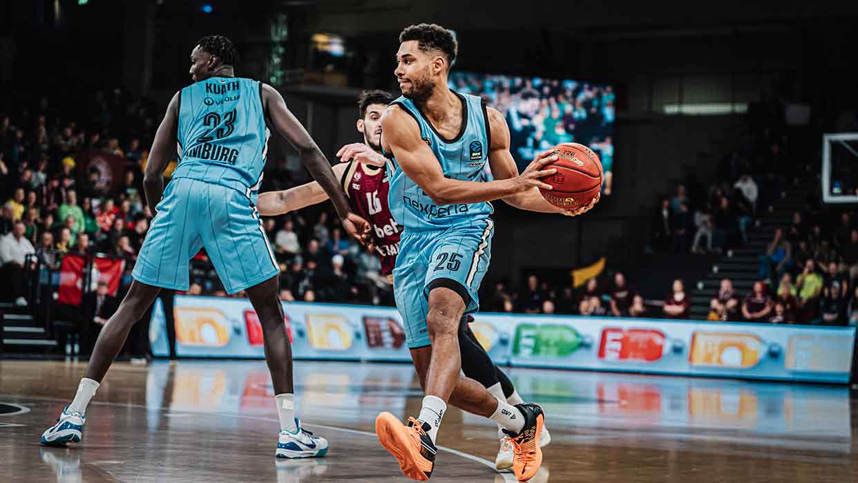 Veolia Towers Hamburg Basketballspieler in hellblauen Trikots, einer hält den Ball und sucht einen Mitspieler, Spielszene in einer Halle mit Publikum.