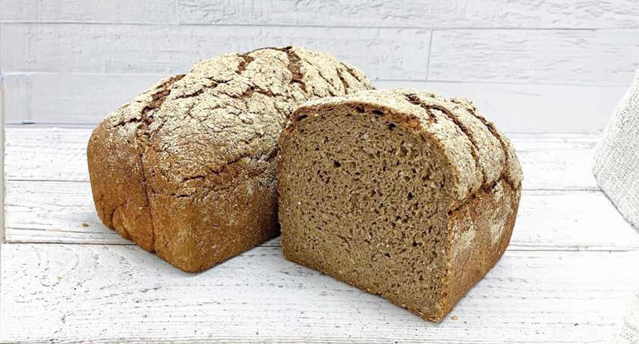 Zwei Laibe Buchweizenbrot, einer ganz und einer halbiert, auf hellem Untergrund vor weißer Wand.