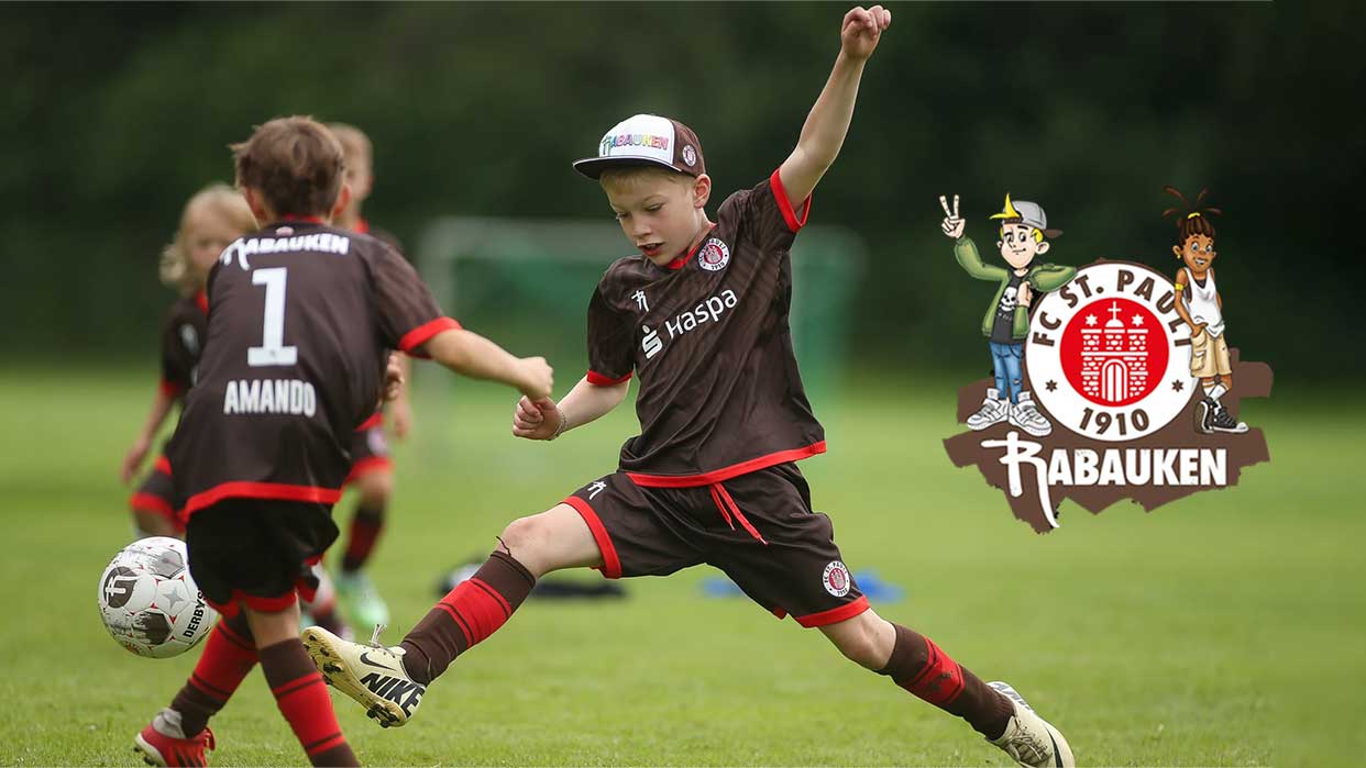 Kinderfußballspieler in schwarzen Trikots mit roten Akzenten spielen auf grünem Spielfeld, ein Spieler schießt den Ball, daneben das Logo von FC St. Pauli Rabauken.