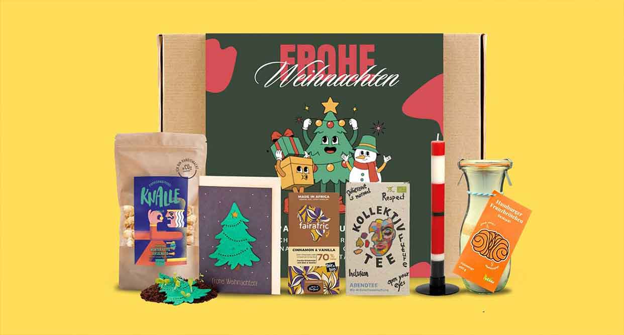 Weihnachtliches Geschenkset mit Schokolade, Tee, Kerze, Saftflasche und bunter Streudeko vor Karton mit Aufschrift 'Frohe Weihnachten'.