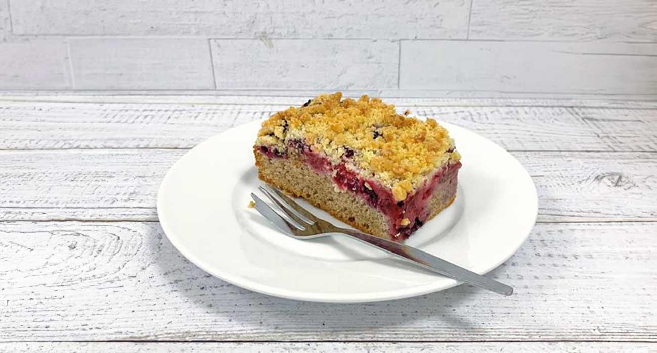 Ein Stück Buchweizenkuchen mit Waldbeeren und Streuseln auf einem weißen Teller mit einer Gabel.